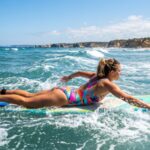 A surfer wearing a colorful polyester swimsuit paddling through ocean waves on a sunny day