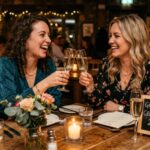 Two women laughing and clinking glasses at a candlelit dinner for Girlfriend Celebration 2026